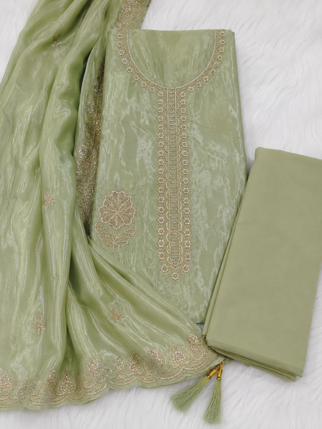 A light sage green ethnic salwar kameez fabric set with gold embroidery, featuring a main kurta, matching dupatta, and folded trouser material on a white textured background.