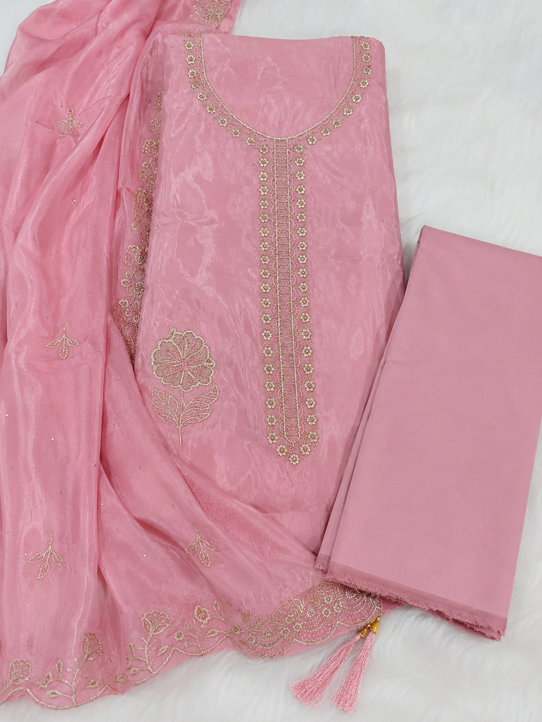 Pink salwar suit material set featuring a printed cotton fabric kurta with gold embroidery, a matching solid pink fabric piece, and a sheer pink dupatta with embroidered borders and tassels, laid out on a white textured background.