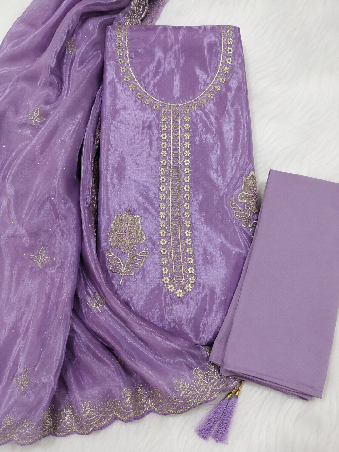 A lavender purple ethnic suit material set featuring a heavily embroidered kurta, a matching sheer dupatta with floral motifs and scalloped border, and a folded matching fabric piece, all displayed on a white textured background.